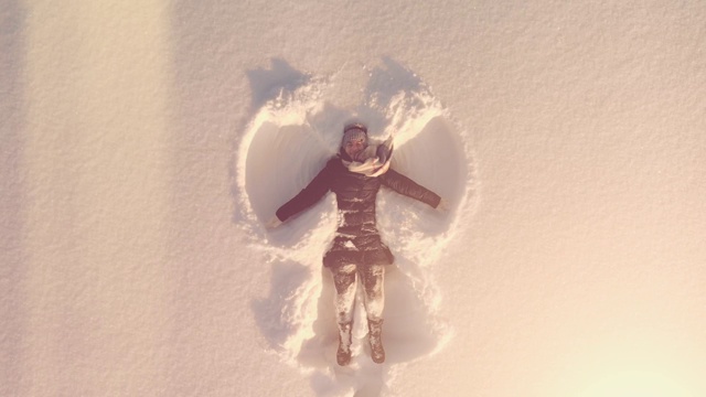 空中俯瞰年轻快乐的女人躺在雪地里，双手和双腿在阳光下做雪天使视频素材