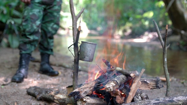 高清慢动作中景拍摄一名身穿迷彩服的士兵在靠近小溪的露营地里用柴火生火在森林里做饭。在营地里燃烧着的火焰。视频素材