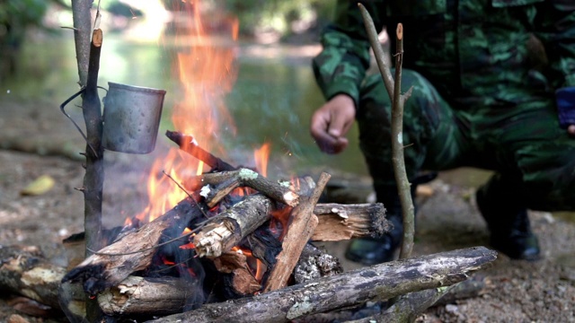 高清慢动作中景拍摄一名身穿迷彩服的士兵在靠近小溪的露营地里用柴火生火在森林里做饭。在营地里燃烧着的火焰。视频素材