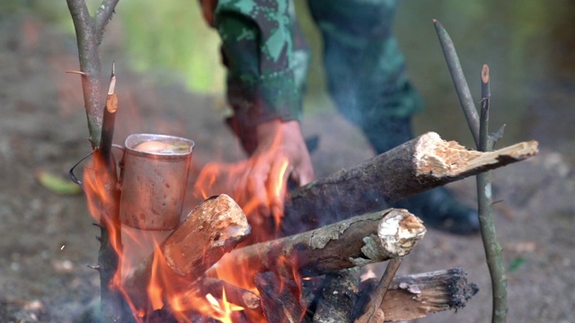 高清慢动作中景拍摄一名身穿迷彩服的士兵在靠近小溪的露营地里用柴火生火在森林里做饭。在营地里燃烧着的火焰。视频素材