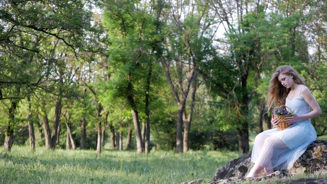 穿着夏装的漂亮女孩坐在森林里的一块岩石上欣赏野花。视频素材