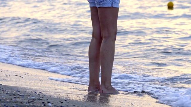 女人光着脚站在夏天的海滩上玩海浪。海浪拍打着她的腿。旅行的概念。视频素材