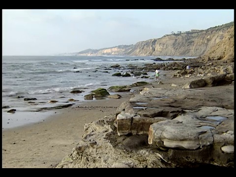 一小群人在海滩上散步时，海浪冲刷着布满岩石的海岸线。视频素材