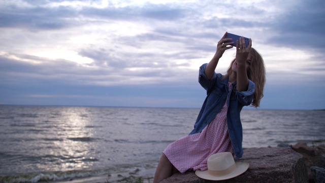 一名年轻女子在海边，在夕阳的背景下在手机上自拍。4 k缓慢莫视频素材