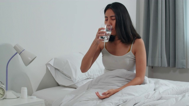 不健康的印度女性遭受失眠。亚洲年轻女子服用止痛药缓解头痛疼痛，喝杯水坐在床上在她的卧室在家里早上。视频素材