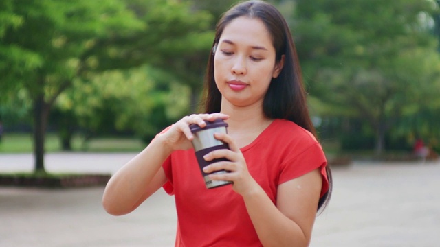 美丽的亚洲年轻女子身穿红色衬衫在城市公园，手拿可重复使用的咖啡杯视频下载