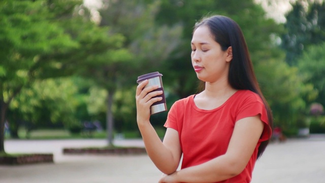 美丽的亚洲年轻女子身穿红色衬衫在城市公园，手拿可重复使用的咖啡杯视频下载