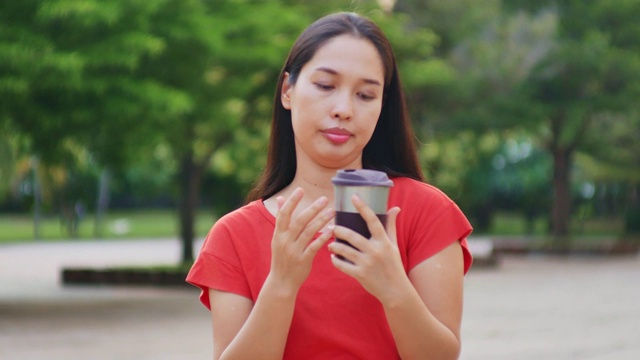 美丽的亚洲年轻女子身穿红色衬衫在城市公园，手拿可重复使用的咖啡杯视频下载