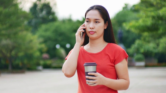 美丽的亚洲年轻女子身穿红色衬衫在城市公园，手拿可重复使用的咖啡杯视频下载