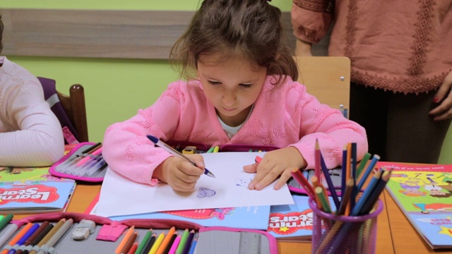 幼儿园学生在课堂上的活动。学龄前儿童在他们的书桌上一起涂色和写作。老师站在他们旁边，给他们提示和鼓励。在托儿所和一群学龄前儿童在一起的老师视频素材