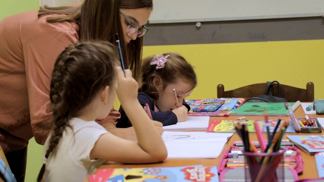 幼儿园学生在课堂上的活动。学龄前儿童在他们的书桌上一起涂色和写作。老师站在他们旁边，给他们提示和鼓励。在托儿所和一群学龄前儿童在一起的老师视频素材