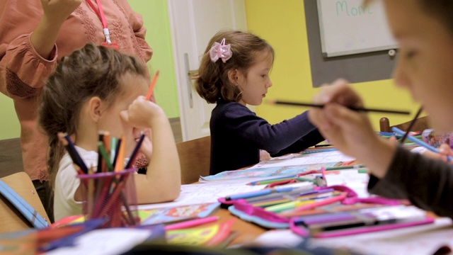 幼儿园学生在课堂上的活动。学龄前儿童在他们的书桌上一起涂色和写作。老师站在他们旁边，给他们提示和鼓励。在托儿所和一群学龄前儿童在一起的老师视频素材