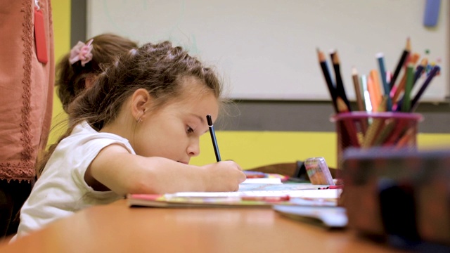 幼儿园学生在课堂上的活动。学龄前儿童在他们的书桌上一起涂色和写作。老师站在他们旁边，给他们提示和鼓励。在托儿所和一群学龄前儿童在一起的老师视频素材