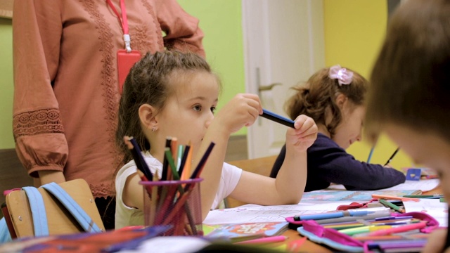 幼儿园学生在课堂上的活动。学龄前儿童在他们的书桌上一起涂色和写作。老师站在他们旁边，给他们提示和鼓励。在托儿所和一群学龄前儿童在一起的老师视频素材