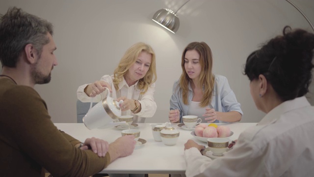 幸福的白人家庭围坐在桌旁，喝茶聊天。成熟的金发女人微笑着把热饮倒进杯子里。不同年龄的人在家里庆祝。视频素材