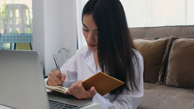 年轻美丽的亚洲女人工作与笔记本电脑和记录坐在家里的客厅，人与技术，在家里工作，在线教育视频素材