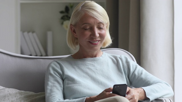 微笑的中年妇女坐在沙发上玩智能手机视频素材