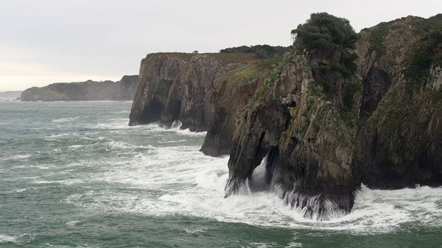 阿斯图里亚斯(Asturias)东海岸的Cueva del Pindal岛(Cueva del Pindal)的悬崖受到海浪的冲击，仿佛想要清除牢牢附着在巨石底部的藤索。该岛有一座天然石拱。视频素材