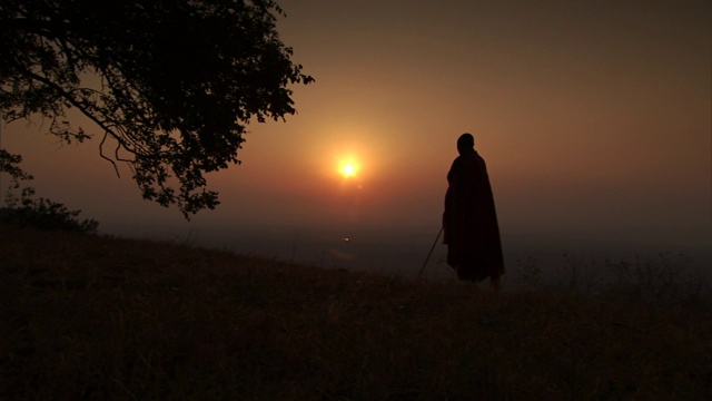 一个马赛人站在长满草的山顶上，金色的天空映出他的轮廓。视频素材