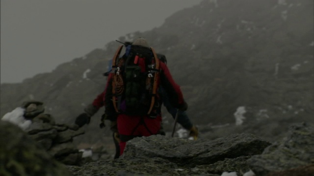 三名成年人背着背包在崎岖的地形上行走，暴风雨在他们周围呼啸而过。视频素材