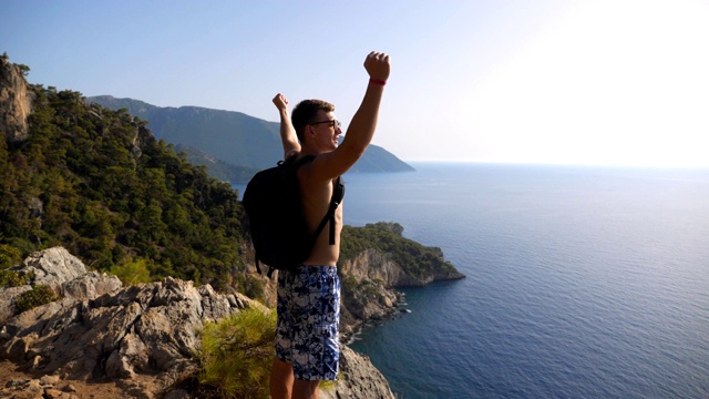 男徒步者站在山顶，胜利地举起双手庆祝成就或成功。人在旅行中积极休息，在山顶享受自由。风景优美的海景背景。多莉拍摄视频素材