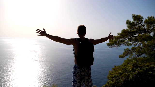 男徒步者背包攀上山顶，举起双手欣赏风景如画的海景。快乐的人在旅途中积极休息，在山顶享受自由。美丽的背景。慢动作视频素材