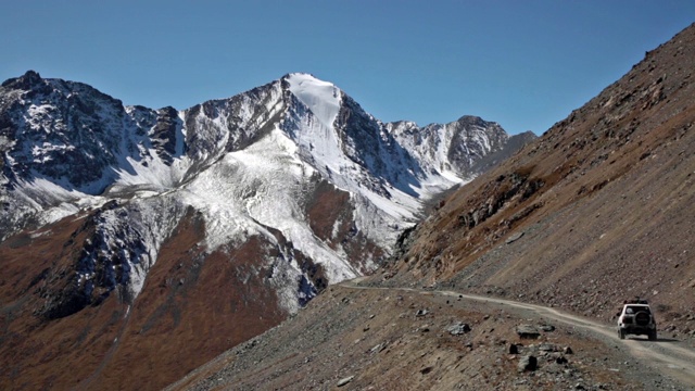 长焦镜头拍摄的白色4x4驾驶在砾石道路通过崎岖的地形和白雪覆盖的雄伟山脉。视频素材