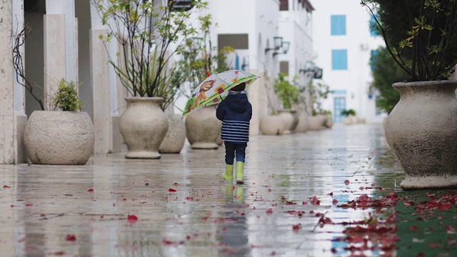 一个穿着胶靴的小男孩撑着伞在雨中走过水坑视频素材