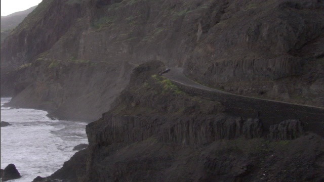 一条公路沿着海洋边一座高大的绿色岩石山的山脚延伸。视频素材