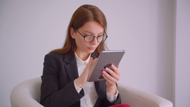 年轻的白人女商人戴着眼镜在平板电脑上发短信的特写，坐在白色公寓的扶手椅上视频素材