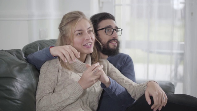 年轻幸福的白人男子拥抱他的金发女友或妻子坐在舒适的沙发上的特写。微笑的家人一起在室内度过周末。视频素材