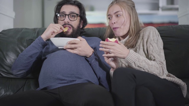 年轻的白人妇女和她怀孕的丈夫一起看电视吃着他盘子里的薯条。有趣的家庭交换社会角色。开玩笑,有趣,开玩笑。视频素材