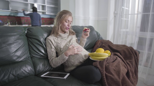 怀孕的白人女孩嚼着苹果，抚摸着自己的肚子。她丈夫站在厨房后面。怀孕，怀孕，健康饮食。视频素材