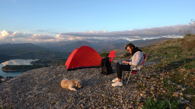 一个年轻人和她的狗在山顶上用笔记本电脑视频素材