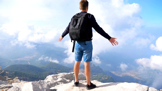 背包徒步者站在山腰，高举双手，享受自由。人们在旅行中积极休息，探索新的地方。游客在山顶欣赏优美的风景。后视图视频素材