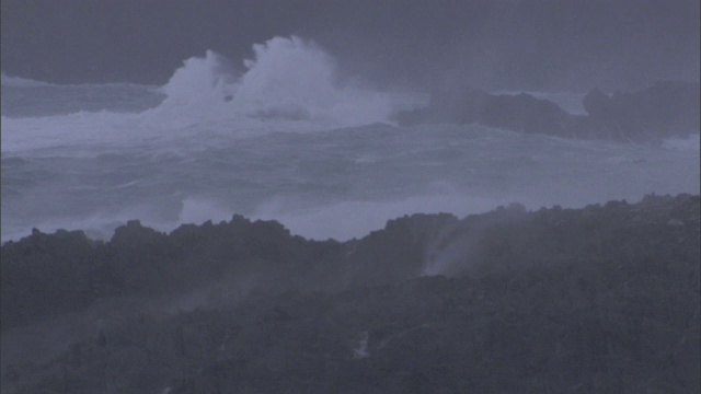 远处的冲浪者冒着暴风雨，汹涌的海浪冲击着岸边的岩石。视频素材