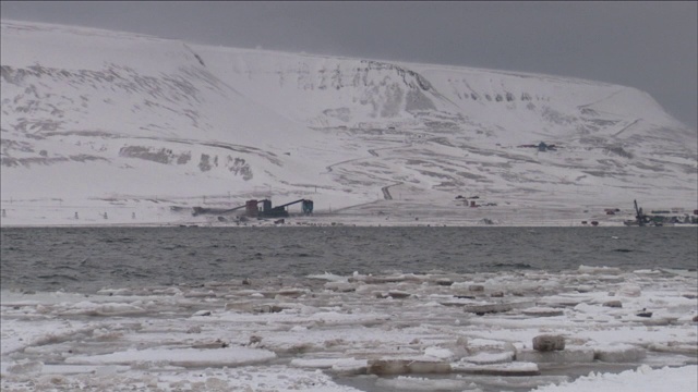 浮冰漂浮在波涛汹涌的水面上，靠近冰雪覆盖的海岸上的工业设备。视频素材