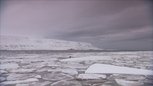 浮冰漂浮在波涛汹涌的水面上，靠近冰雪覆盖的海岸上的工业设备。视频素材