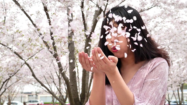 美丽的中国年轻女子在樱花盛开的森林里，穿着粉红色的连衣裙，用手吹着粉红色的樱花花瓣，动作超级慢。视频素材