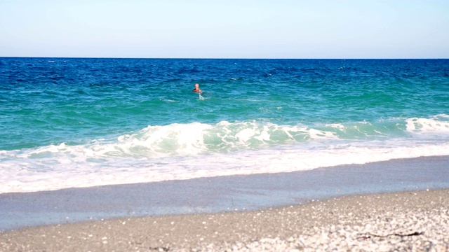 在蓝绿色的海水里，有两个人在游泳。夏天炎热的晴天。海浪在海上，在沙滩上。带着白色泡沫的波浪被冲上岸视频素材