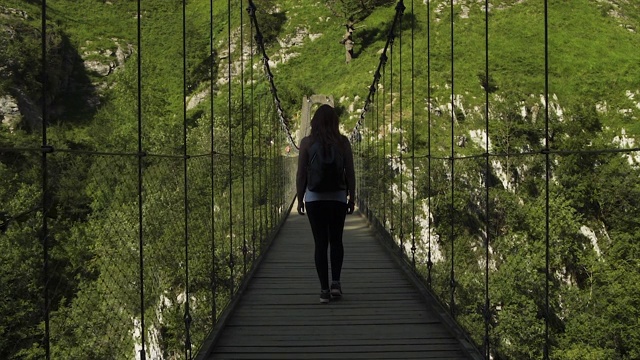 女探险家红发女孩行走，徒步旅行，穿过美丽的霍尔扎特人行桥，木制金属灰色桥，黑色背包，绿树森林，山在夏季的慢镜头4K。视频素材