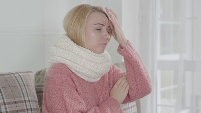 年轻迷人的白人妇女在白色围巾颤抖的肖像。金发女孩坐在家里的沙发上打喷嚏。医疗保健医疗疾病传染病视频素材