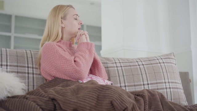 一个悲伤的白人妇女坐在家里的沙发上，用手捂着脸。等待丈夫或男友的沮丧女孩。关系问题,心烦意乱的。视频素材