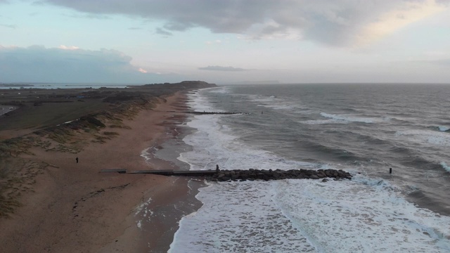 在暴风雨的灰色天空下，海浪在海滩上的冲击镜头通过一个岩石的防波堤(防波堤)视频素材