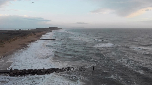 在暴风雨的灰色天空下，海浪在海滩上的冲击镜头通过一个岩石的防波堤(防波堤)视频素材