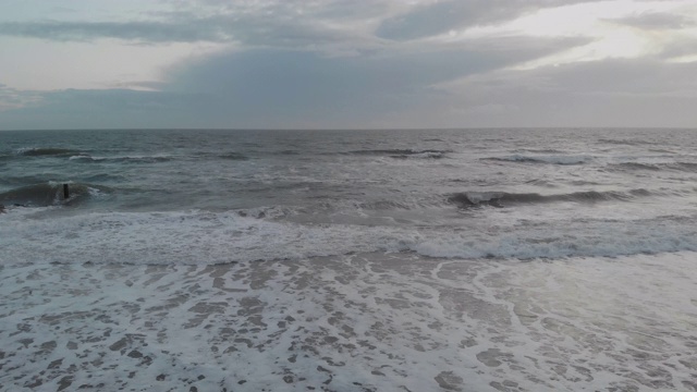 一个空中缓慢的侧边(从右到左)的镜头，在暴风雨的灰色天空下，冲击海滩的海浪通过一个岩石的防波堤视频素材