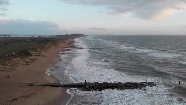 在暴风雨的灰色天空下，海浪在海滩上的冲击镜头通过一个岩石的防波堤(防波堤)视频素材