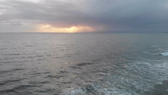 一个空中向前的镜头，在一个暴风雨的灰色天空下的波涛汹涌的海与太阳在云后发光视频素材