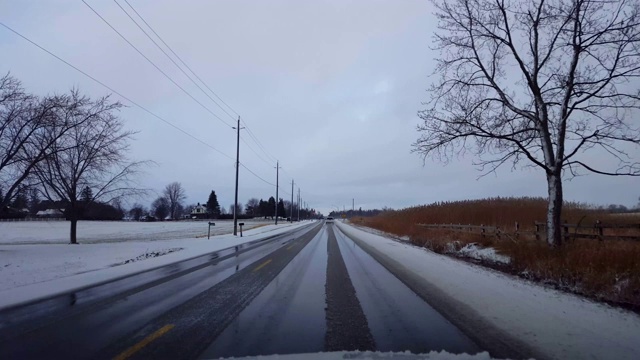 驾驶在冬天的乡村道路与雪在道路的一边在白天。司机的观点POV驾驶街道与雪在树上与阴云天空。视频素材