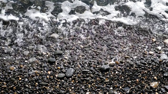 在阳光明媚的日子里，小浪拍打着卵石滩。海浪冲刷着多石的海岸。潮湿的石头在明亮的阳光下闪闪发光。美丽的海景。旅行的概念。慢镜头视频素材